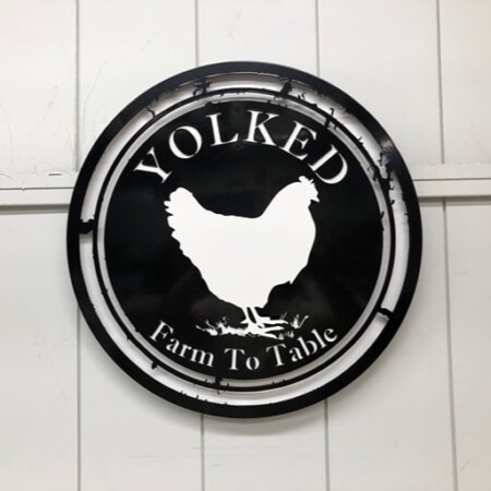 Yolked Farm To Table round sign, a white hen cut against a black disc inside a raw steel ring.