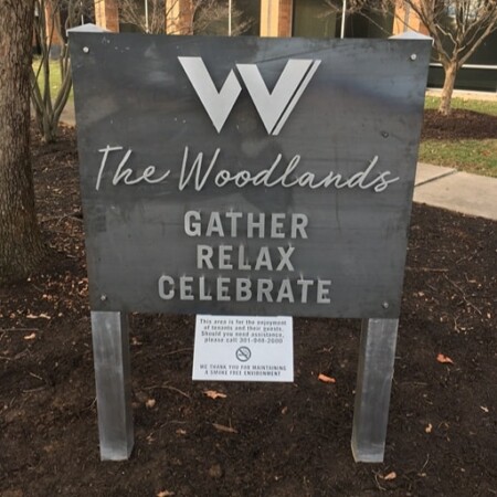 The Woodlands courtyard sign in mottled steel reading Gather, Relax, Celebrate, post-mounted in a mulched bed.