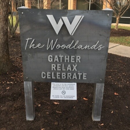 The Woodlands steel courtyard sign with the office building behind it, its W monogram cut above the lettering.