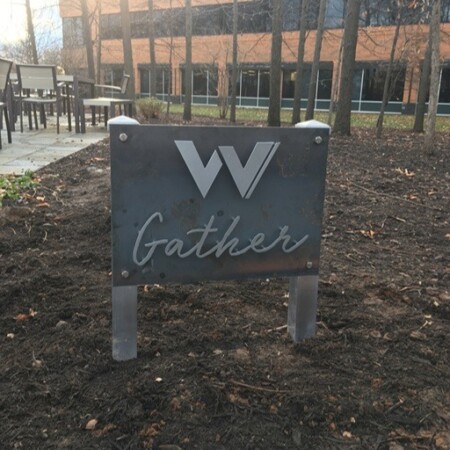 Gather marker in mottled steel with a W monogram, post-mounted beside the courtyard seating.