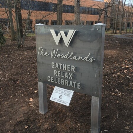 The Woodlands steel sign viewed from the path, bare trees and the office block behind it.
