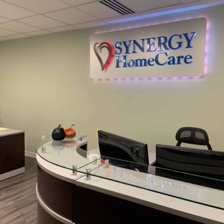 Synergy HomeCare reception sign lit above the front desk, the name in red and blue on a white panel.