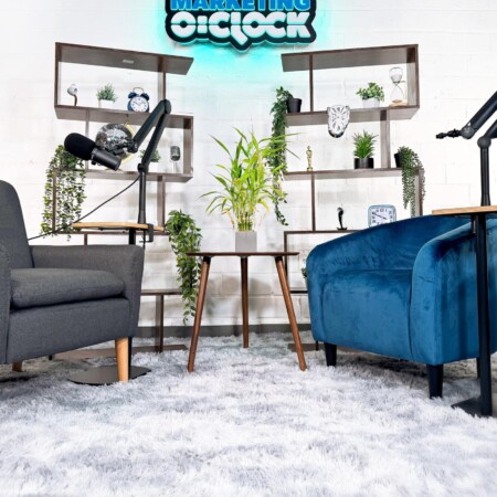 Marketing O'Clock sign lit on the wall of the podcast studio, above the chairs and shelving.